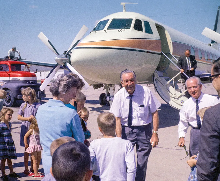 Walt Disney, an senior man in a white button up short-sleeved shirt, with a tie and slacks, greets a small crowd of people in front of his airplane, which is large enough it extends out of frame and is white with orange and blue stripes.