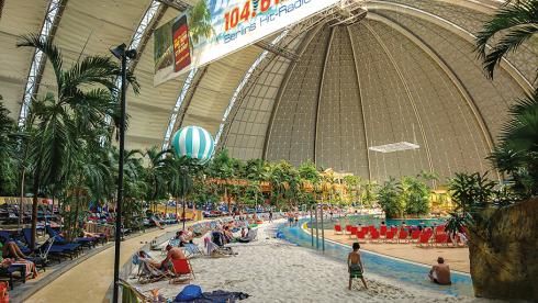 A beachfront resort with green tropical trees and sunbathers. sits inside the dome of the Cargolifter hangar.