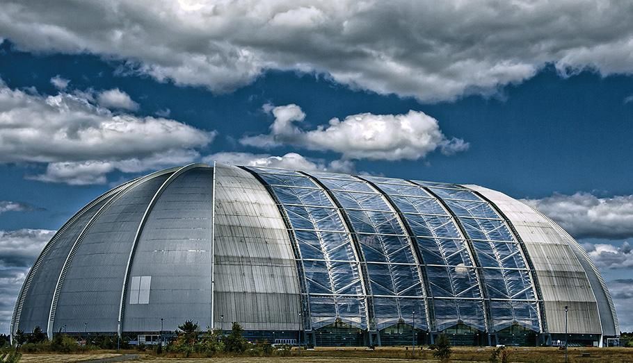 A huge hangar, covered in silver-reflective material, sits framed by an intense blue, cloud-filled sky in the background.
