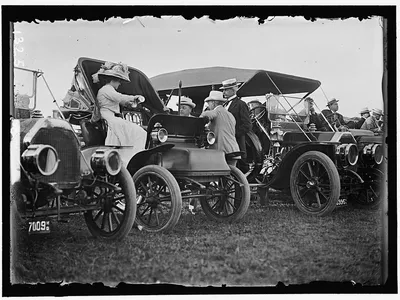 A black and white photograph featuring cars with people conversing.