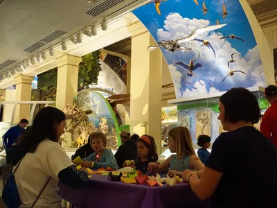 A group of children and two adults sit at a table creating origami birds among exhibits.