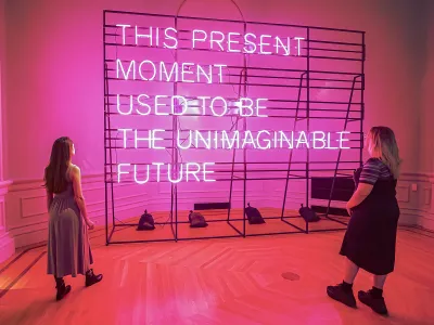 Two people stand in front of a pink neon sign that states "This Present Moment Used to Be the Unimaginable Future"