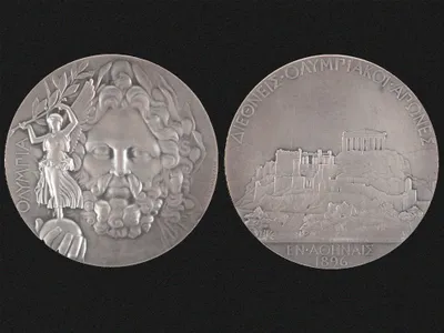One side of the silver medal depicts Zeus holding Nike in the palm of his hand, while the other side features the Acropolis in Athens.
